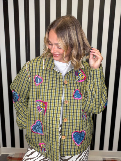 A woman wearing a green and yellow plaid button-up jacket decorated with colorful embroidered hearts, shown against a black and white striped background.