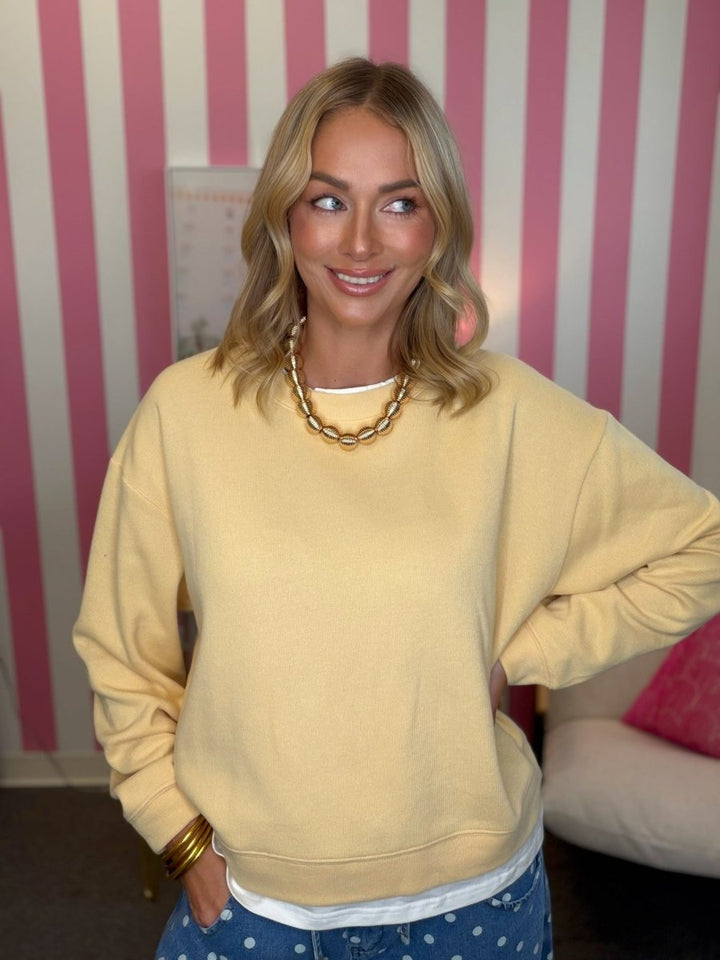 A woman wearing a pale yellow oversized crewneck sweatshirt, blue polka dot jeans, and a chunky gold chain necklace, standing in front of a pink and white striped wall.
