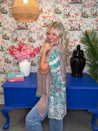 A woman modeling a two-piece linen set consisting of a black top and a matching long vest in a bohemian-themed room with floral wallpaper, a blue table, and a woven lamp.