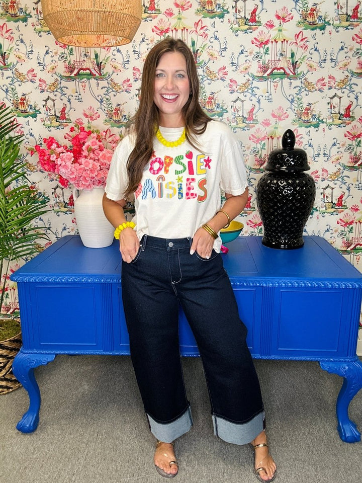 Woman wearing a colorful t-shirt and jeans sitting on a blue table with floral and decorative wall background