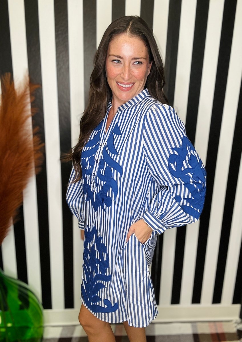 A woman modeling a blue and white vertically striped shirt dress with blue abstract ikat patterns on the front and sleeves, standing in front of a black and white striped wall.