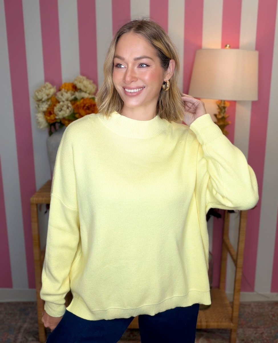 A woman wearing a pale yellow crewneck sweatshirt and dark blue pants, standing in front of a pink and white striped wall with a lamp and flowers in the background.