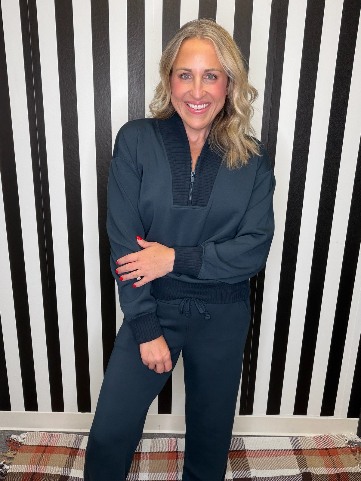 A woman wearing a matching dark teal lounge set consisting of a quarter-zip sweatshirt and drawstring sweatpants, standing in front of a black and white striped wall.