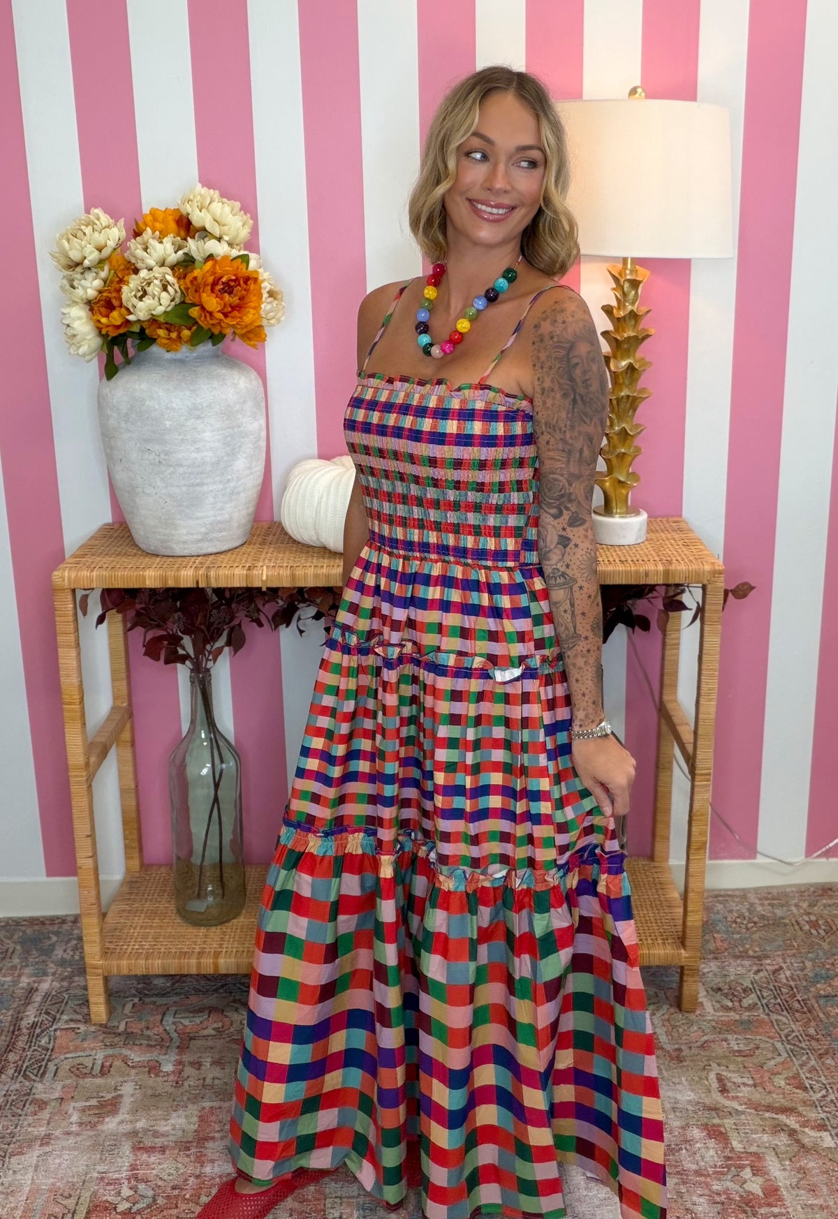 A woman modeling a colorful plaid halter maxi dress with tiered ruffles, standing in front of a pink and white striped wall with a wooden console table and flowers.