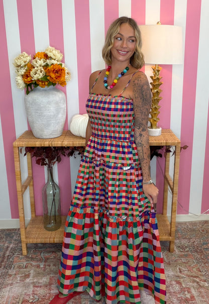 A woman modeling a colorful plaid halter maxi dress with tiered ruffles, standing in front of a pink and white striped wall with a wooden console table and flowers.