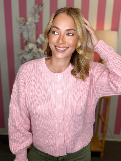 A woman wearing a pink chunky knit button-up cardigan sweater, standing in front of a pink and white striped wall.