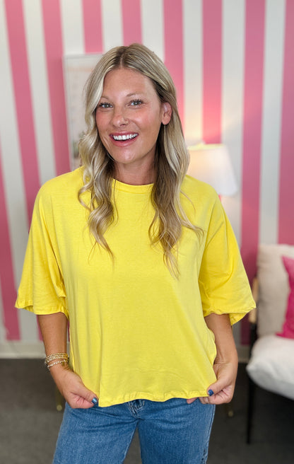 A smiling blonde woman wearing a bright yellow short-sleeve t-shirt and blue jeans, standing in front of a pink and white striped wall.