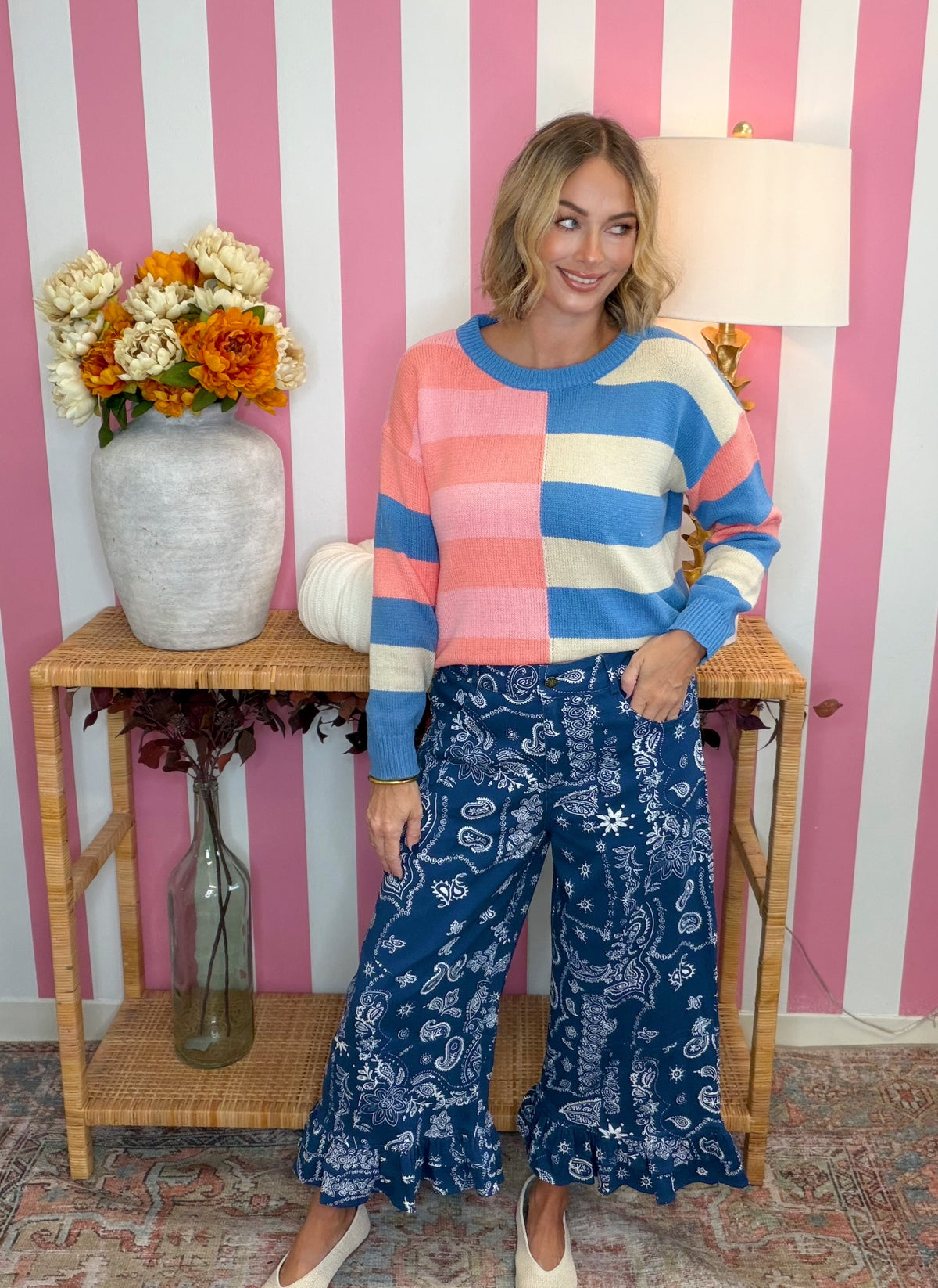 A woman modeling a colorful striped knit sweater and blue paisley wide-leg pants, standing in front of a pink and white striped wall with a side table and flowers.
