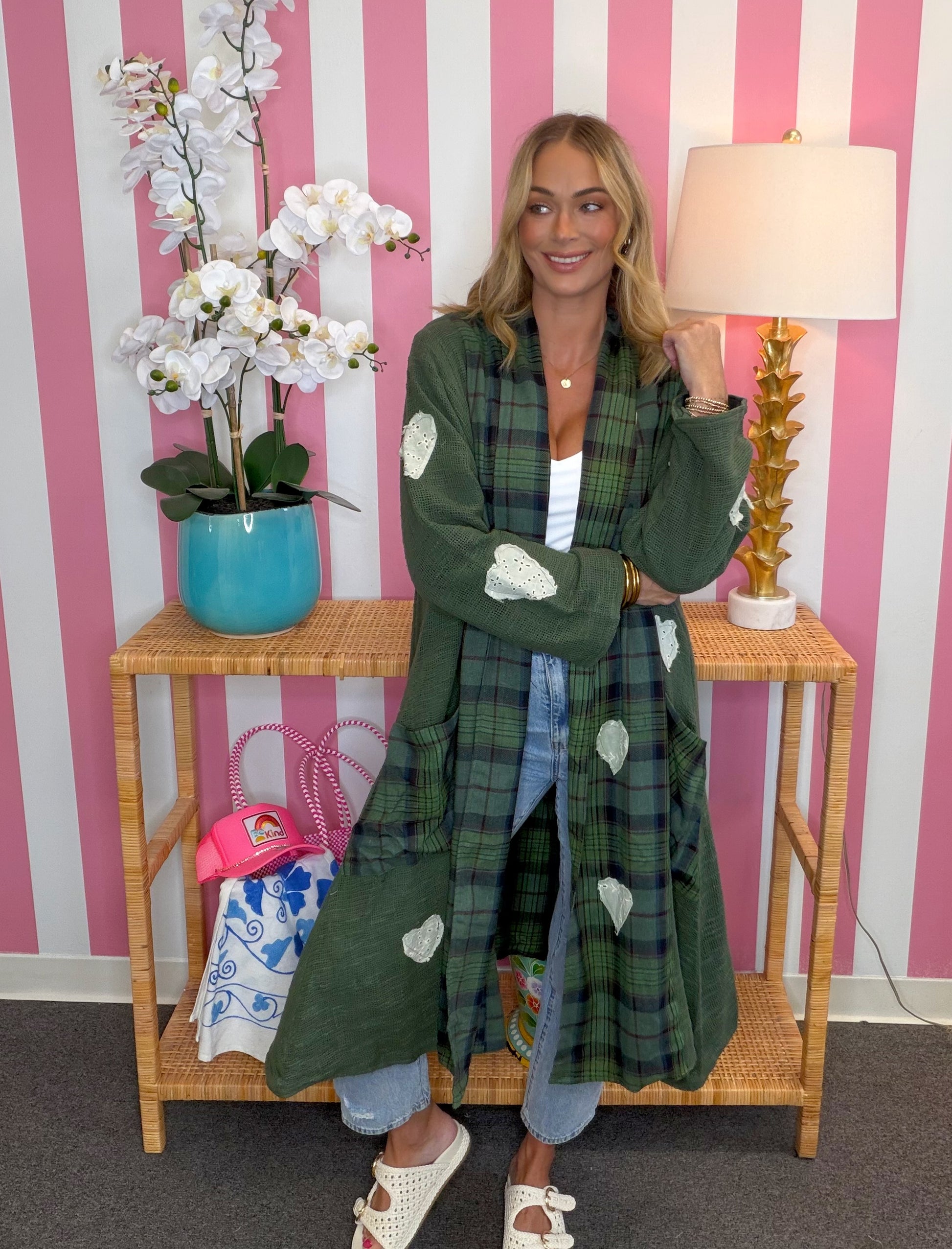 A woman models a green plaid long coat with white patchwork details, standing in front of a pink and white striped wall next to a wooden side table with a lamp and flowers.