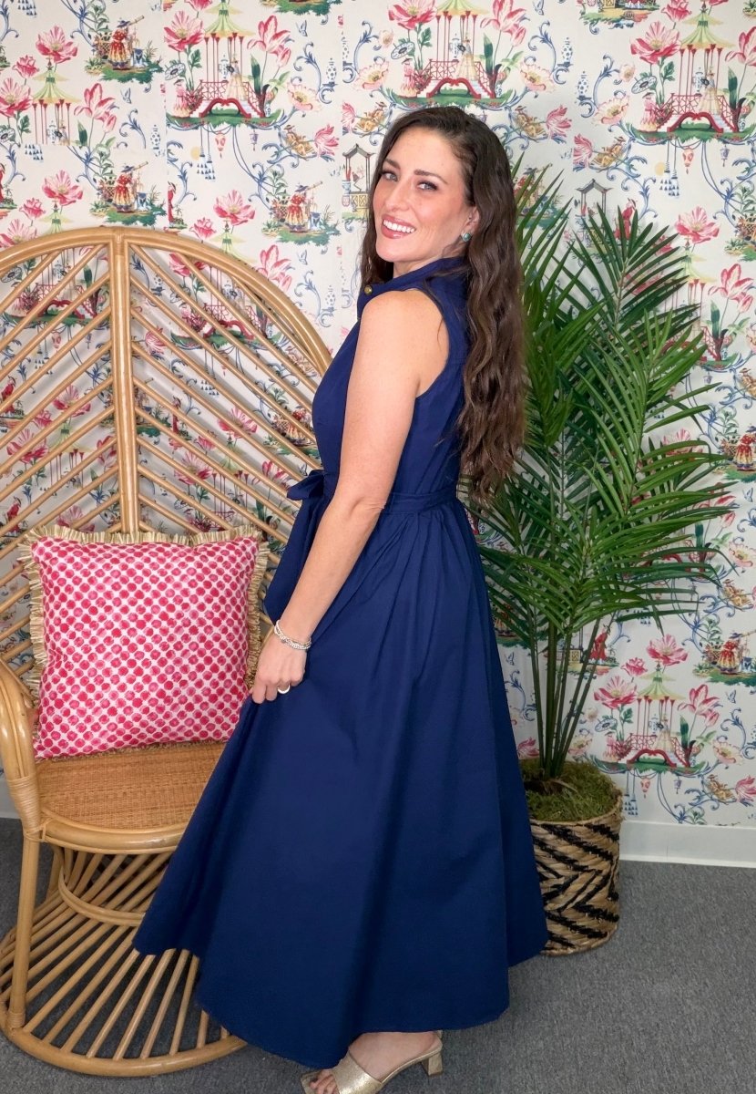 Woman in a blue dress standing in a room with floral wallpaper and a wicker chair.