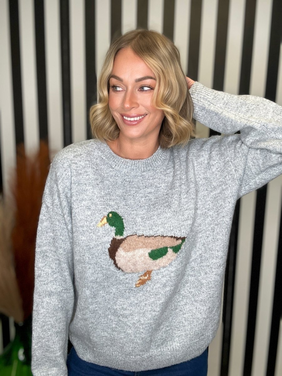 A woman wearing a light gray knit sweater with a large embroidered mallard duck on the front, posed against a black and white striped background.