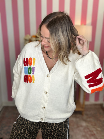 A woman wearing a cream-colored knit cardigan with colorful 'HO! HO! HO!' text on the chest and a red-and-orange striped pattern on the sleeve, paired with leopard-print pants, set against a pink and white striped wall background.