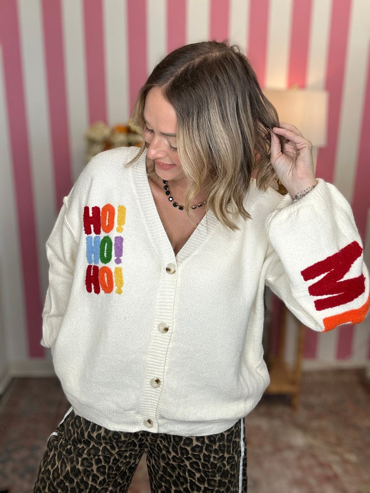 A woman wearing a cream-colored knit cardigan with colorful 'HO! HO! HO!' text on the chest and a red-and-orange striped pattern on the sleeve, paired with leopard-print pants, set against a pink and white striped wall background.