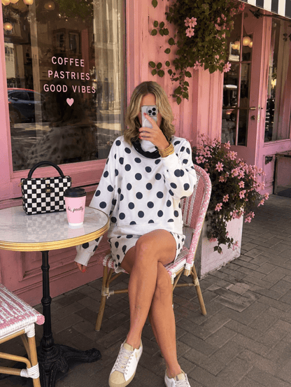 A woman takes a mirror selfie while sitting on a pink wicker chair outside a pink cafe. She is wearing a white knit sweater and matching mini skirt with black polka dots, white sneakers, and holding a pink coffee cup next to a black-and-white checkered handbag.