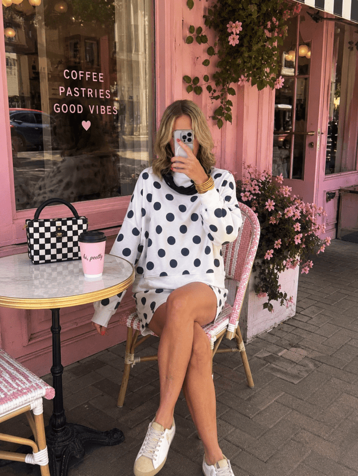 A woman takes a mirror selfie while sitting on a pink wicker chair outside a pink cafe. She is wearing a white knit sweater and matching mini skirt with black polka dots, white sneakers, and holding a pink coffee cup next to a black-and-white checkered handbag.