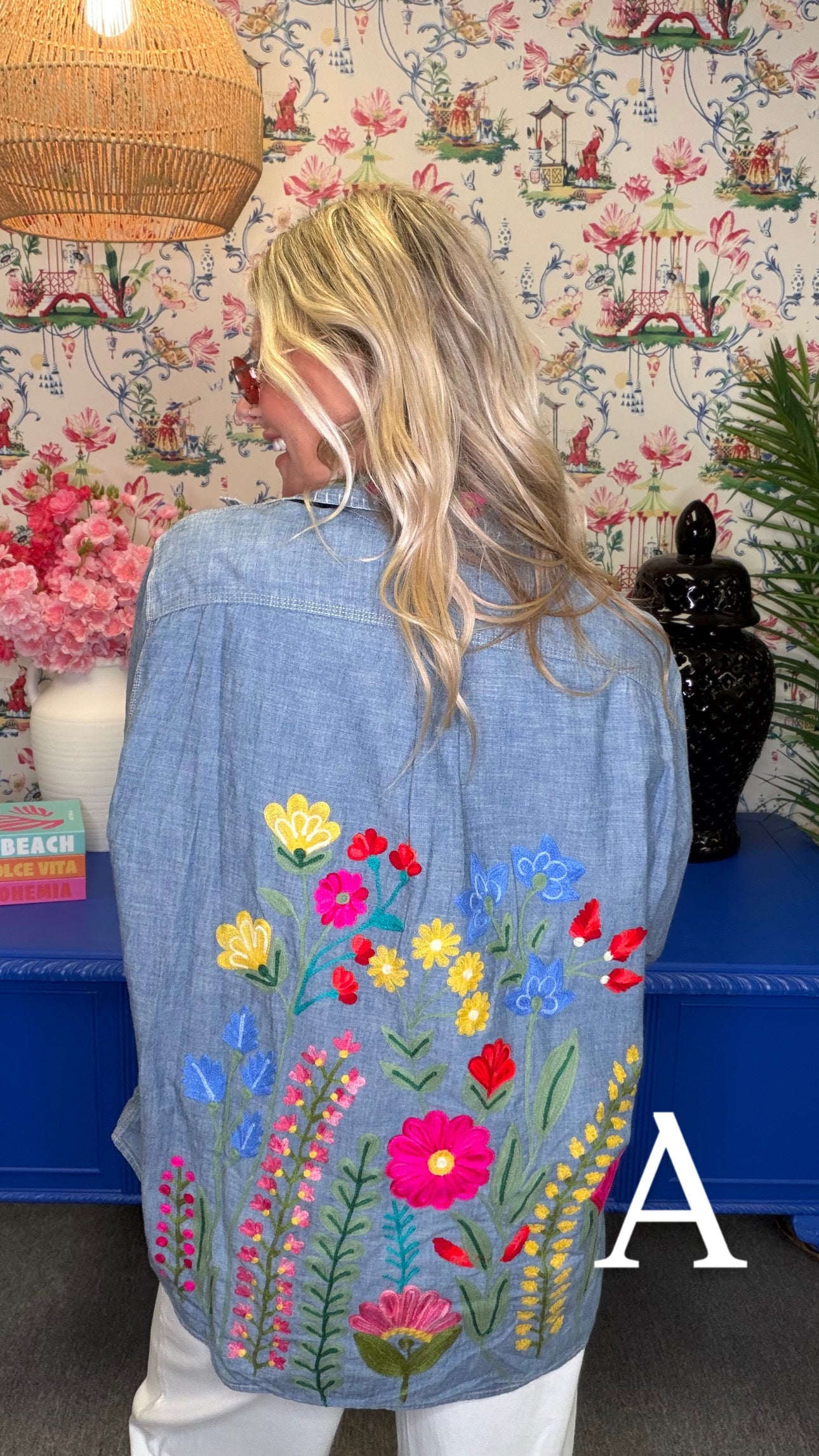 A woman viewed from behind wearing a light blue denim shirt jacket with colorful embroidered wildflowers on the back, set against a patterned wallpaper background.