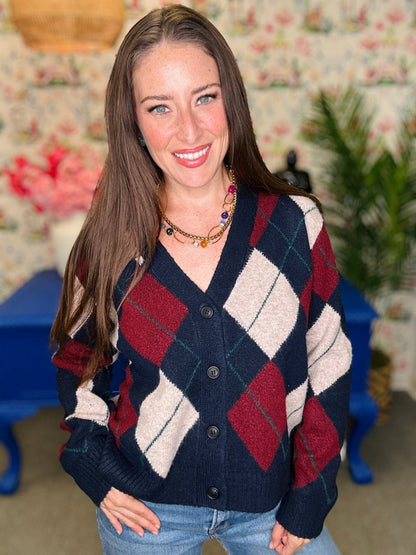 A woman smiling and wearing a navy blue, burgundy, and cream argyle knit cardigan sweater with blue jeans, standing in a vintage-style room with floral wallpaper and blue furniture.