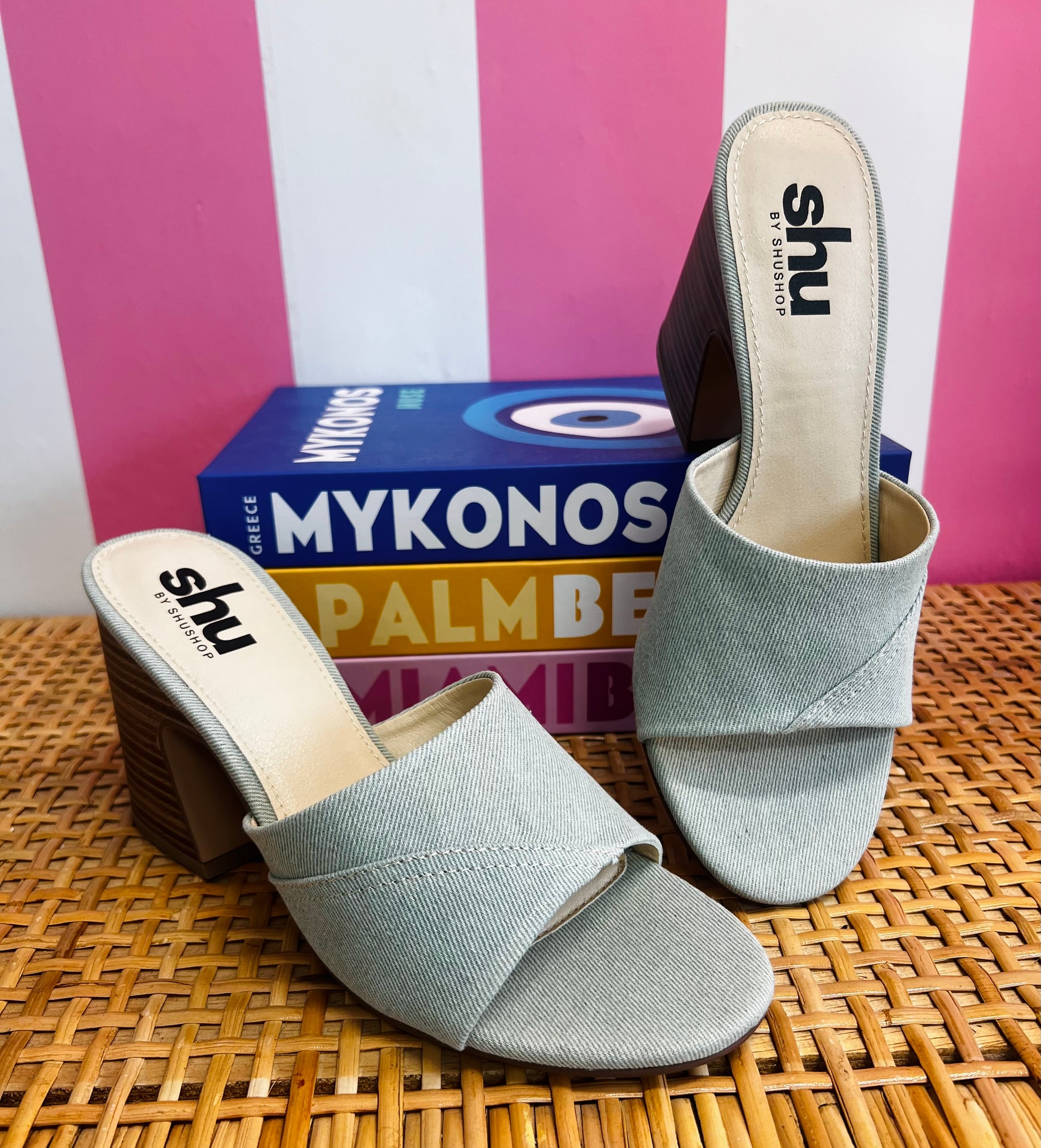 Pair of light blue open-toe sandals with 'shu' branding on a woven surface with a striped pink and white background.
