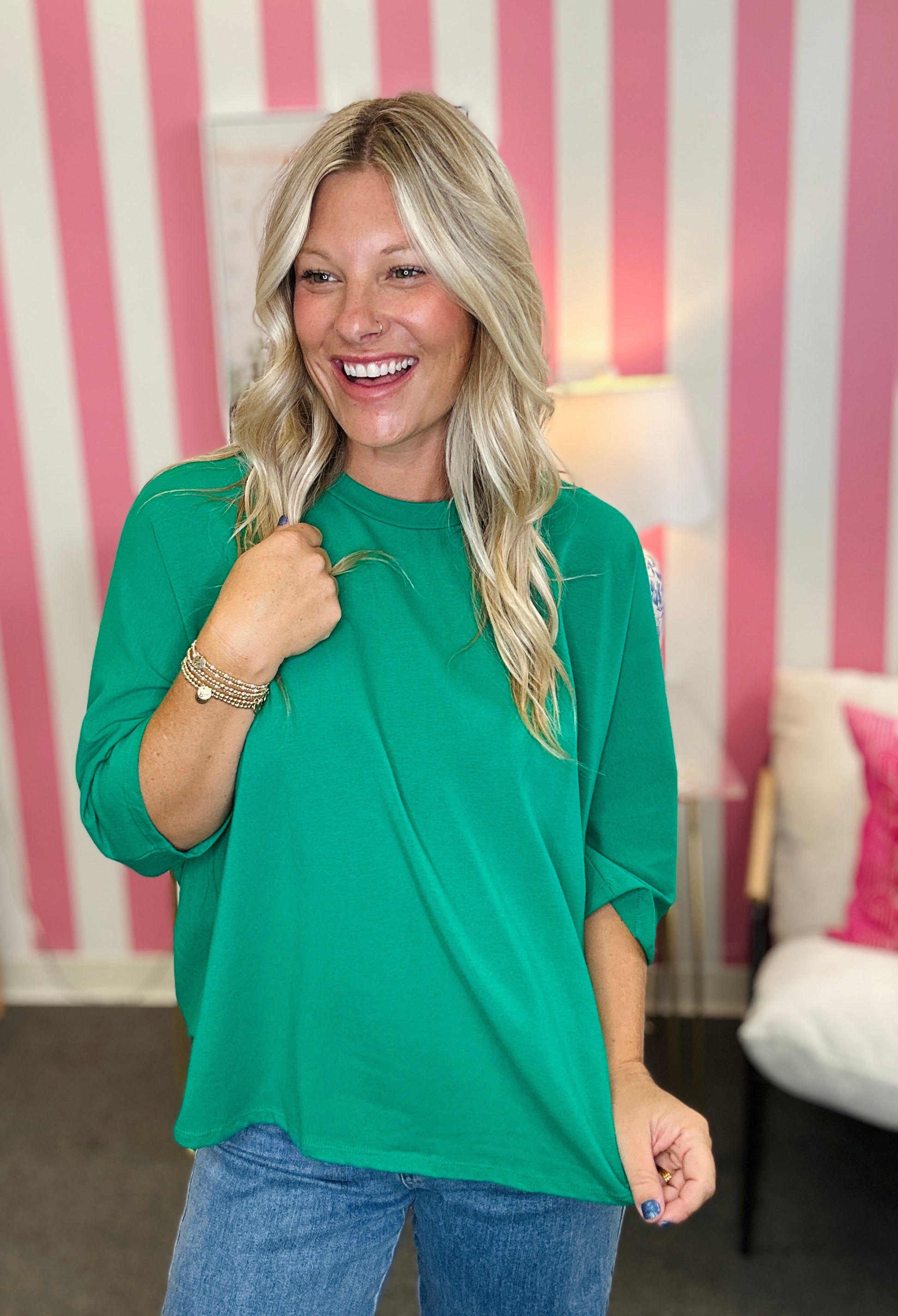 A smiling blonde woman wearing a bright green oversized three-quarter sleeve t-shirt and blue jeans, standing in front of a pink and white striped wall.