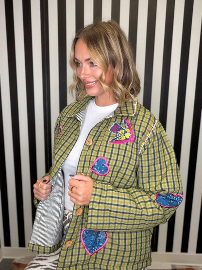 A woman wearing a green and yellow plaid quilted jacket with colorful sequined heart patches, styled over a white t-shirt, standing against a black and white striped wall.