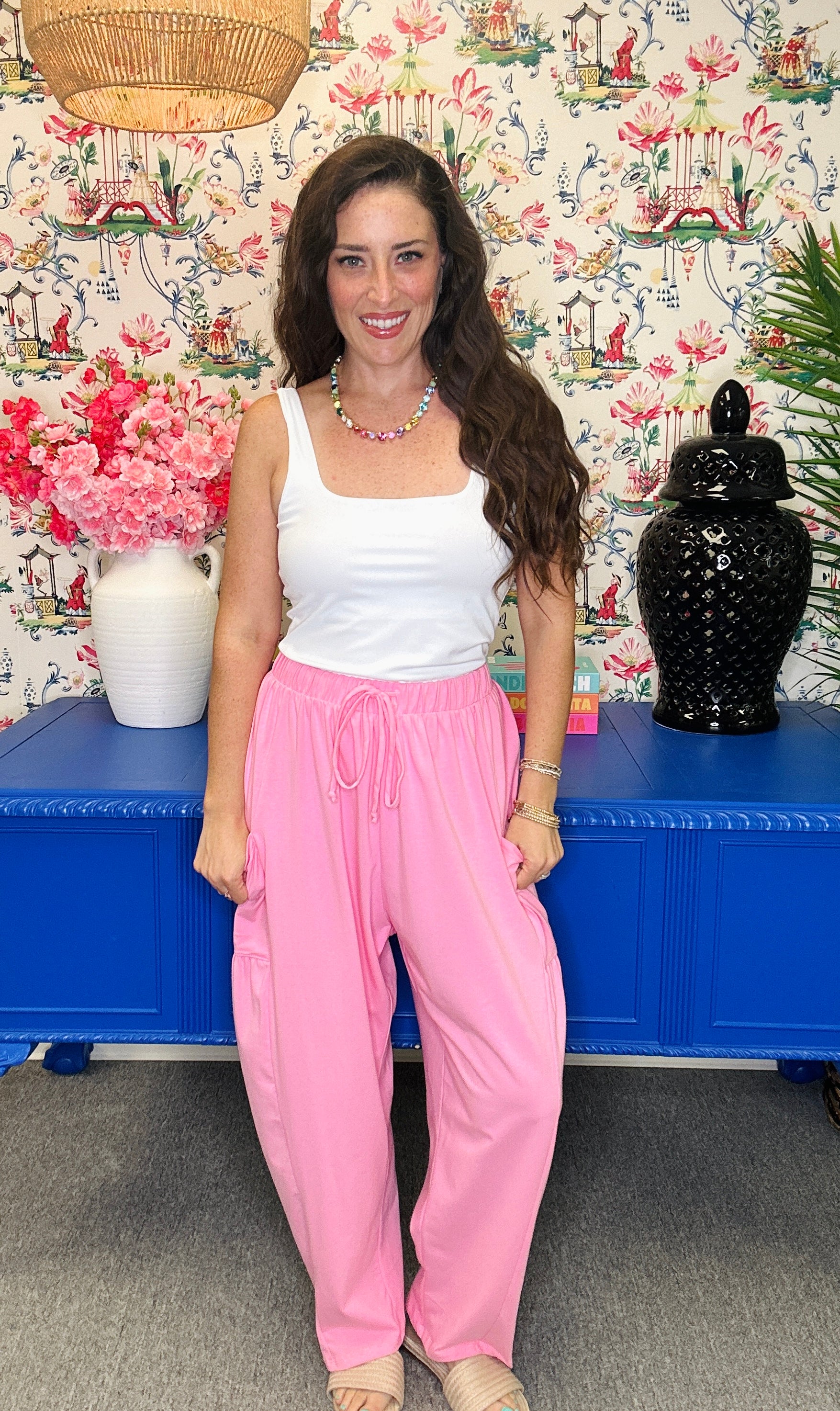 A woman stands indoors wearing a white sleeveless tank top and bright pink drawstring pants. She is posed in front of a blue cabinet, floral wallpaper, a vase of pink flowers, and a black decorative jar.