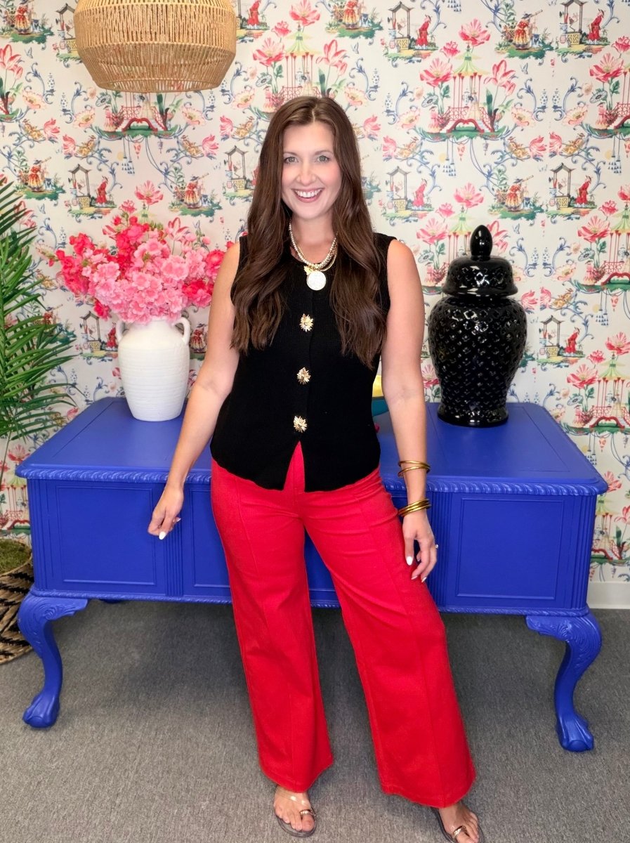 Woman in black top and red pants standing in front of a blue table with floral wallpaper.