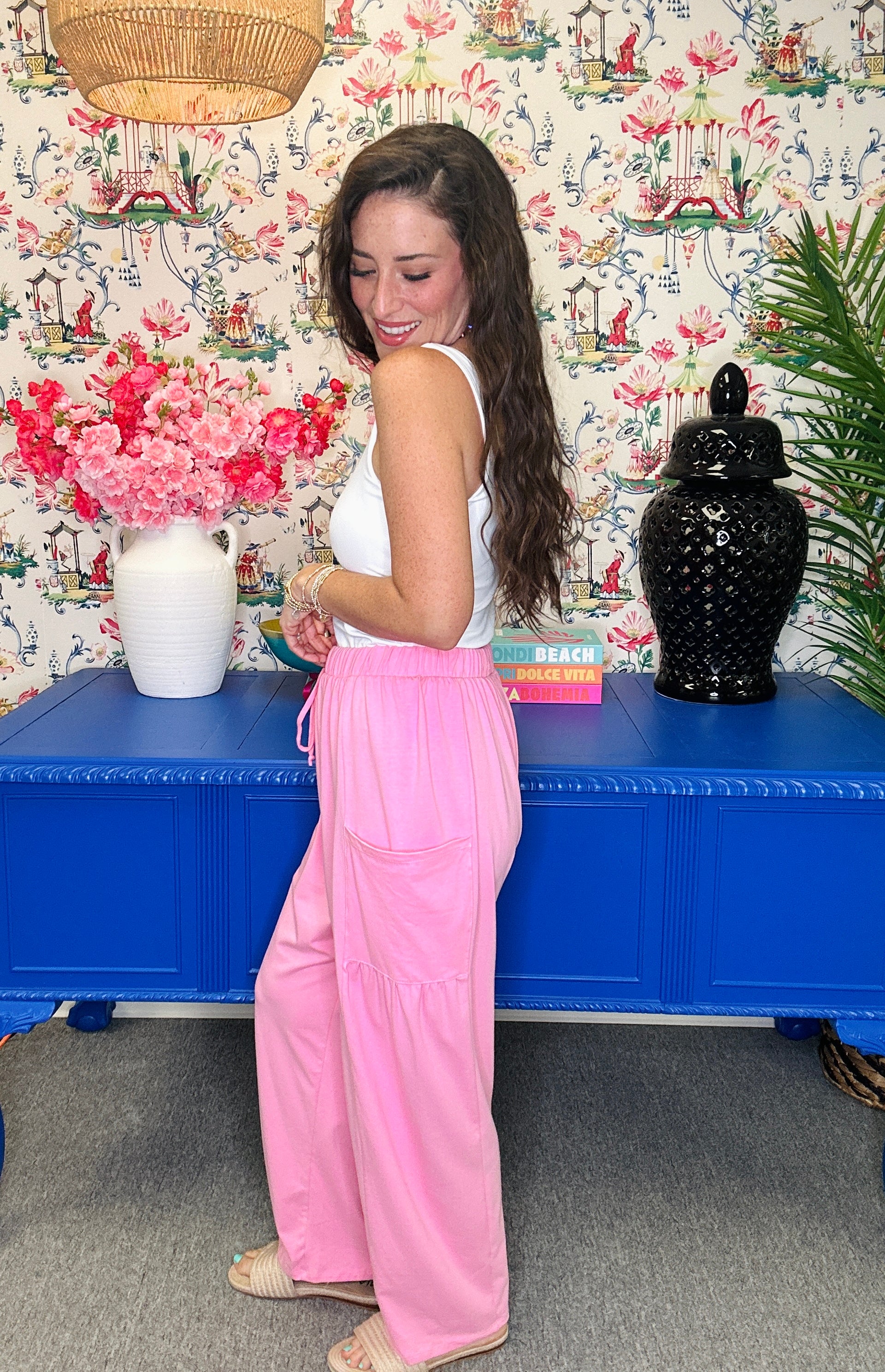 A woman wearing a white sleeveless top and pink wide-leg pants stands in front of a blue cabinet in a decorated room with floral wallpaper, a vase of pink flowers, and a black decorative jar.