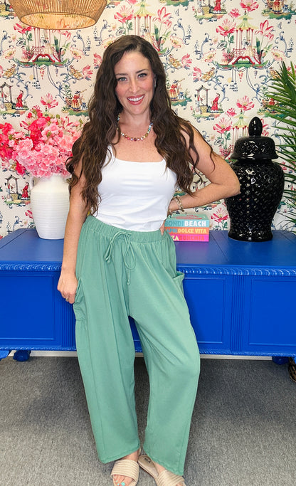 A woman stands indoors wearing a white tank top and light sage green wide-leg drawstring pants, posed in front of a floral wallpaper wall and a bright blue cabinet.