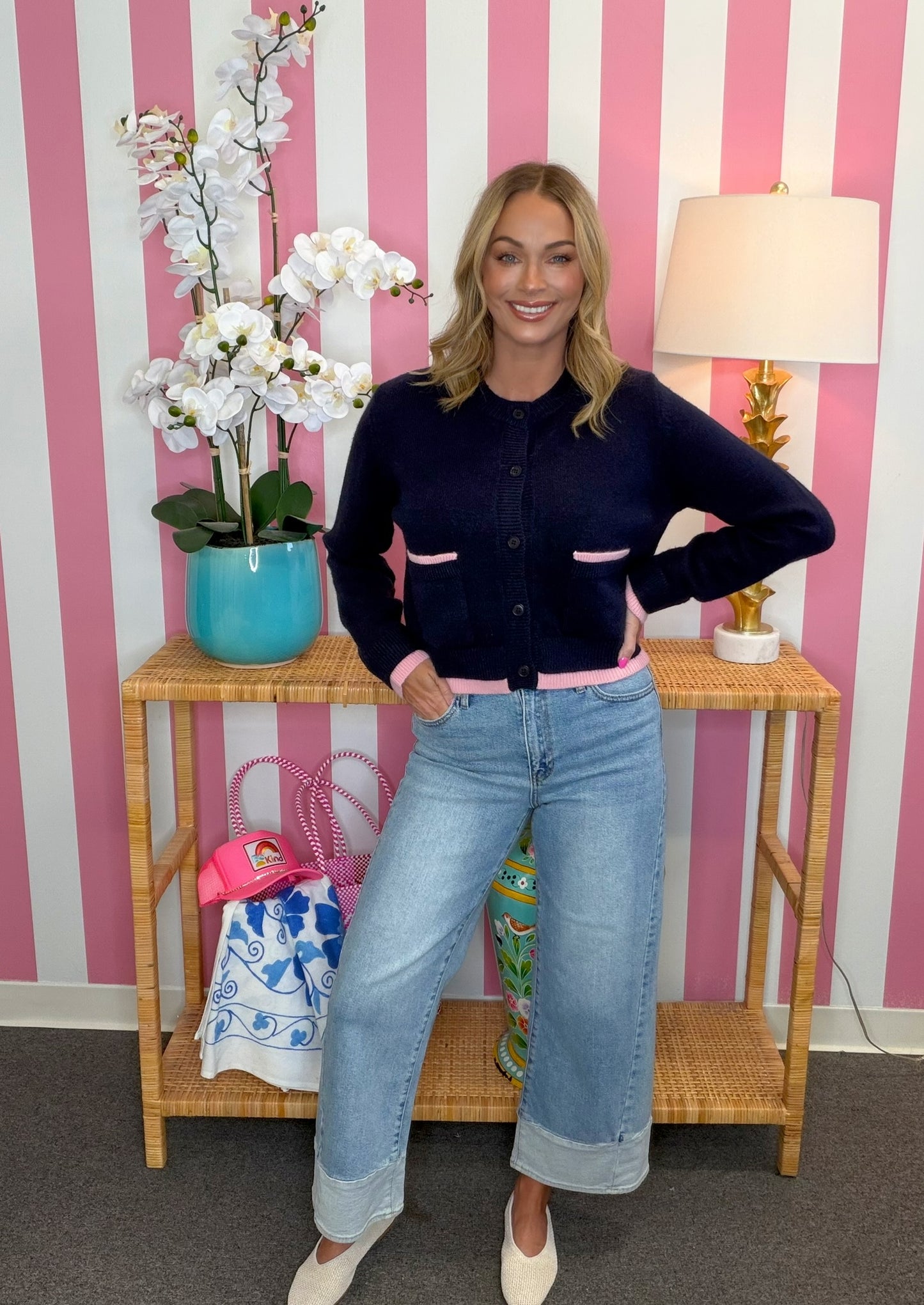 A woman modeling a dark navy blue cardigan with pink pocket trim and a pink hem, paired with light-wash blue jeans and cream-colored shoes, standing in front of a pink and white striped wall.