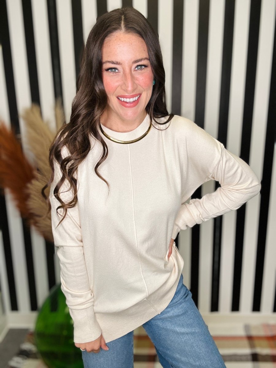 A woman with long wavy brown hair smiling, wearing a cream-colored long-sleeve knit sweater with a gold collar trim and blue jeans, standing in front of a black and white striped background.