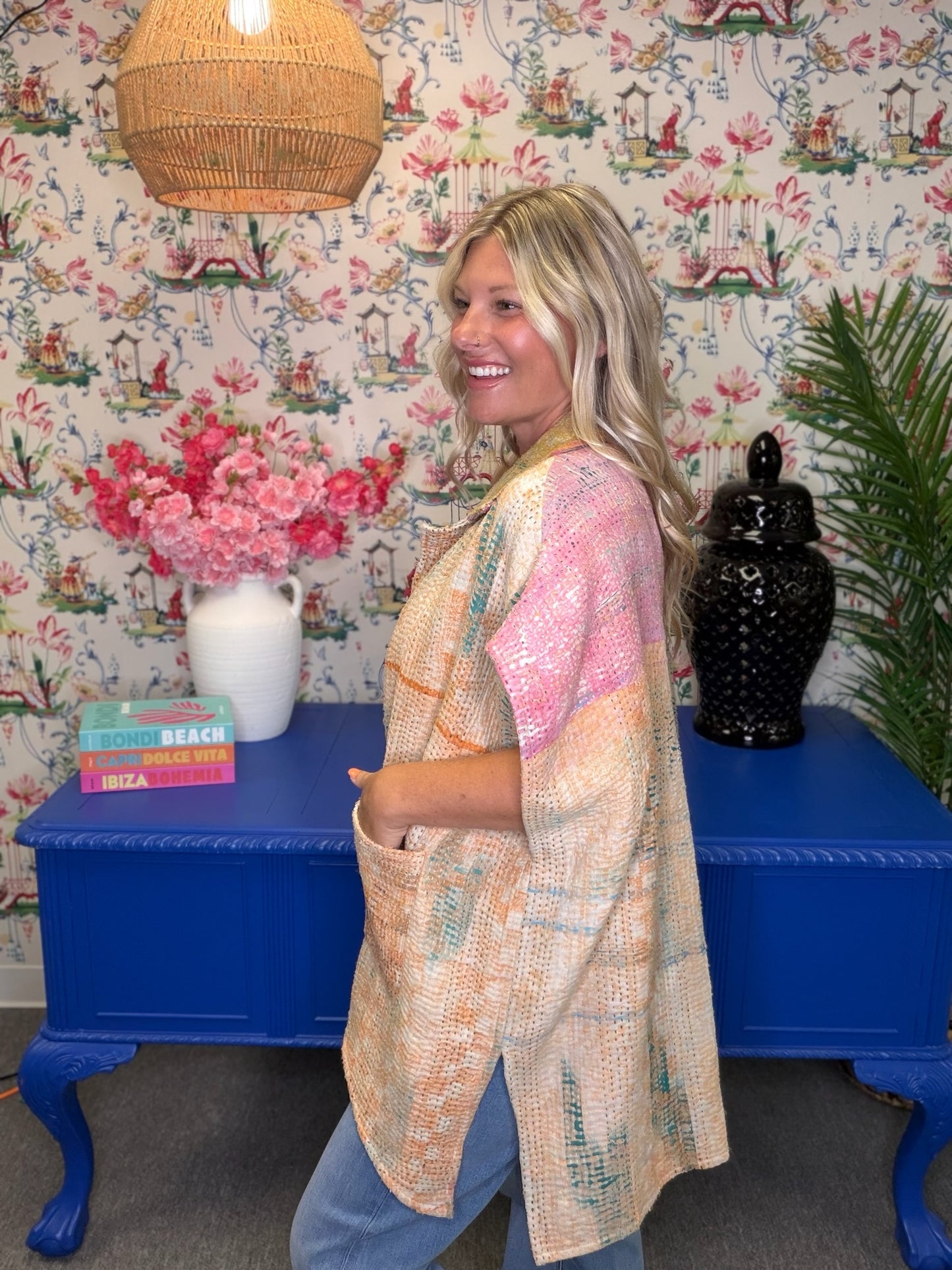 A woman wearing a lightweight, open-front poncho-style top with a multicolor pastel pattern, standing in a decorated room with floral wallpaper, a blue side table, and a wicker lamp.