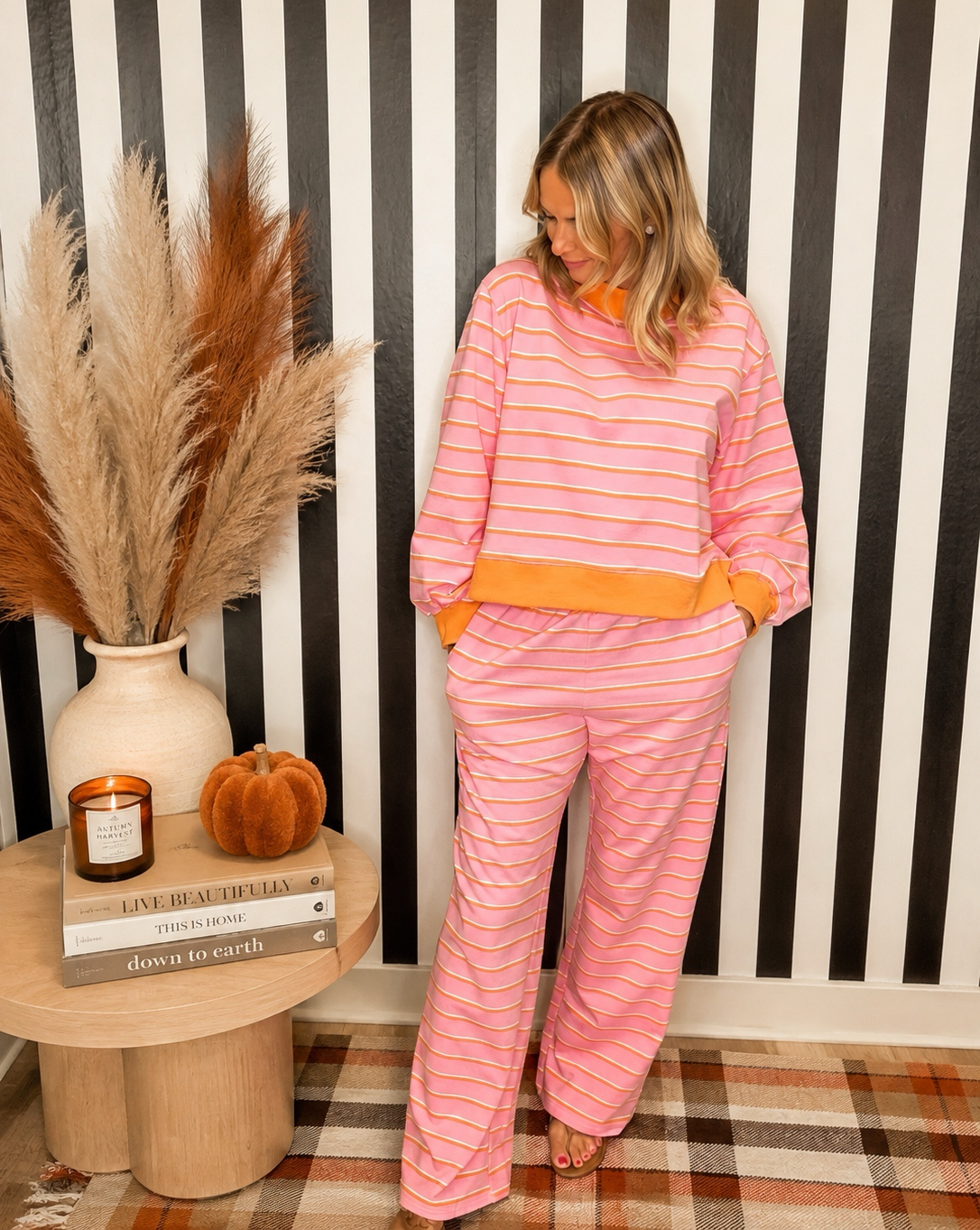 A woman models a pink and white striped lounge set consisting of a long-sleeve top and matching pants, styled in a home setting with a black and white striped wall, a wooden side table with pampas grass, and a plaid rug.