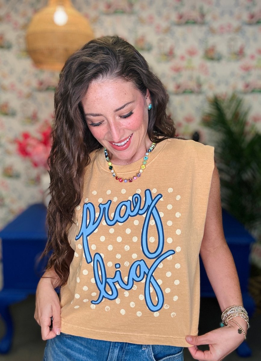 A woman wearing a sleeveless tan top with white polka dots and blue cursive text reading 'pray big' on the front, paired with blue jeans.