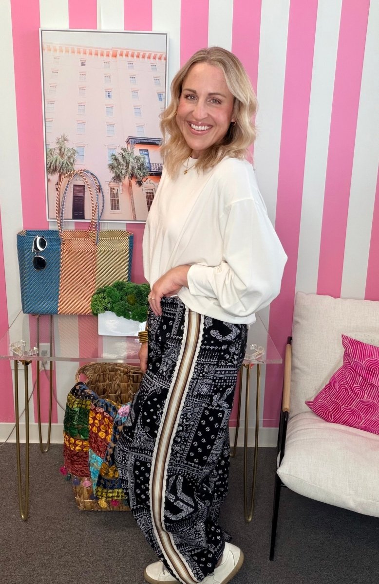 A woman stands in a pink and white striped room wearing a cream long-sleeve blouse and black wide-leg pants with a white stripe and bandana print. She is smiling and posing next to a glass table with handbags and a white chair.