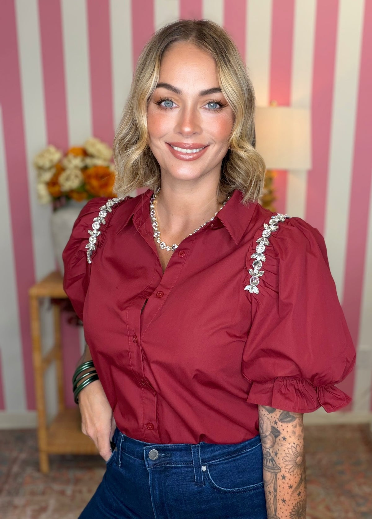 A woman wearing a burgundy puff-sleeve button-up blouse with white beaded trim along the shoulders and collar, paired with blue jeans, standing in front of a pink and white striped wall.