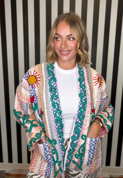 A woman wearing a colorful bohemian-style embroidered cardigan over a white t-shirt, standing in front of a black and white striped background.