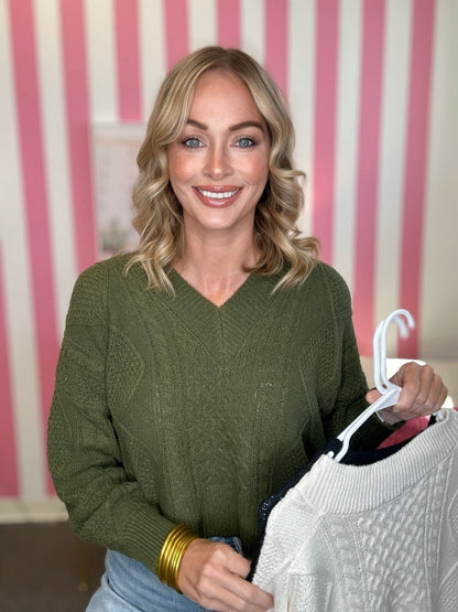 A smiling blonde woman wearing an olive green cable-knit sweater, holding up a white cable-knit sweater on a hanger against a pink and white striped background.