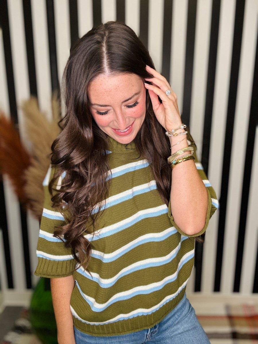 A woman with long wavy brown hair wearing a short-sleeve knit sweater with horizontal olive green, white, and light blue stripes, paired with blue jeans. She is smiling and looking down, with her hand near her hair, set against a black and white striped background.