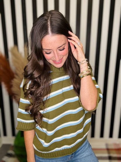 A woman with long wavy brown hair wearing a short-sleeve knit sweater with horizontal olive green, white, and light blue stripes, paired with blue jeans. She is smiling and looking down, with her hand near her hair, set against a black and white striped background.