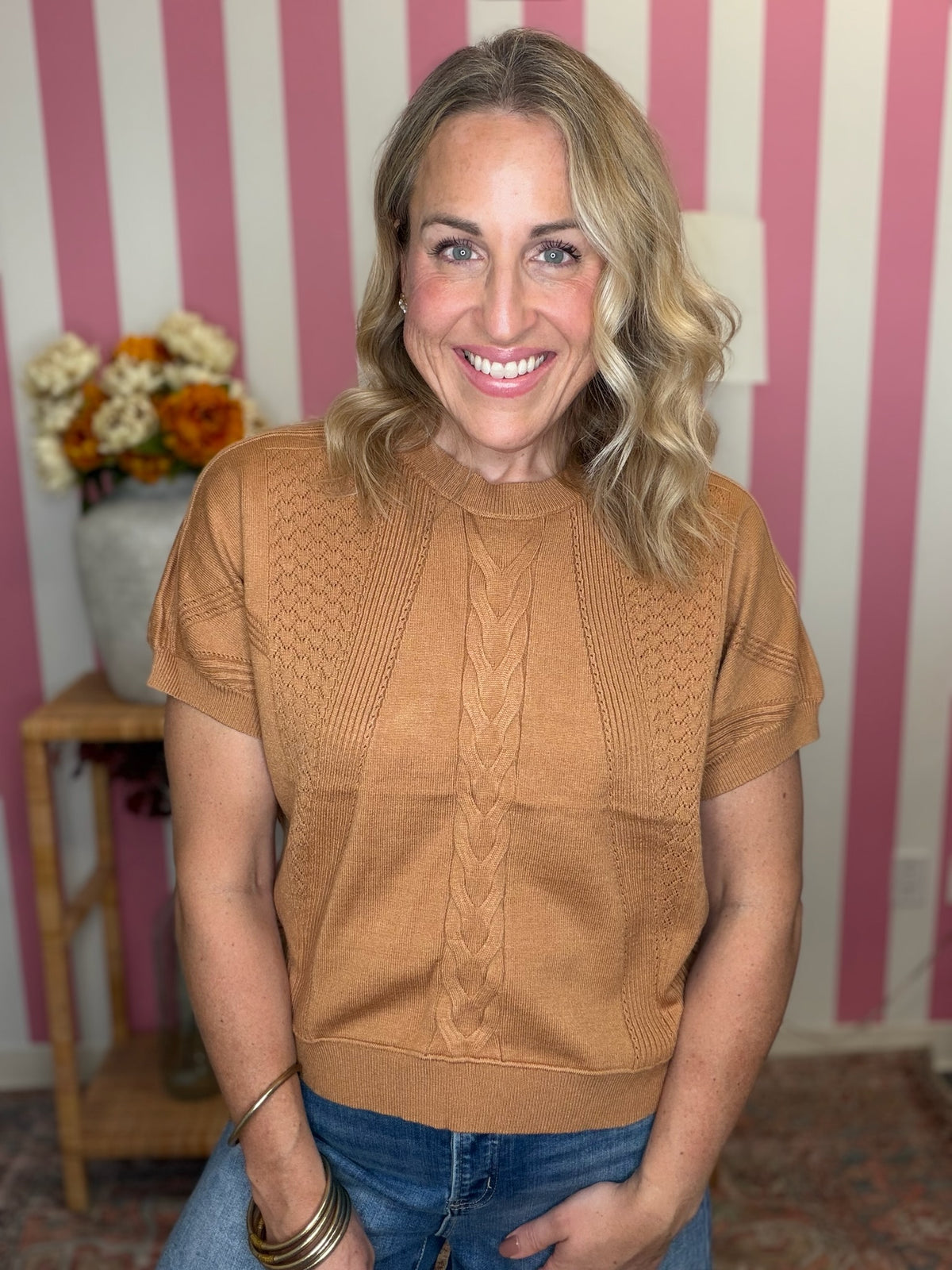A smiling blonde woman wearing a tan short-sleeve knit sweater with cable and textured patterns, paired with blue jeans, standing in front of a pink and white striped wall.