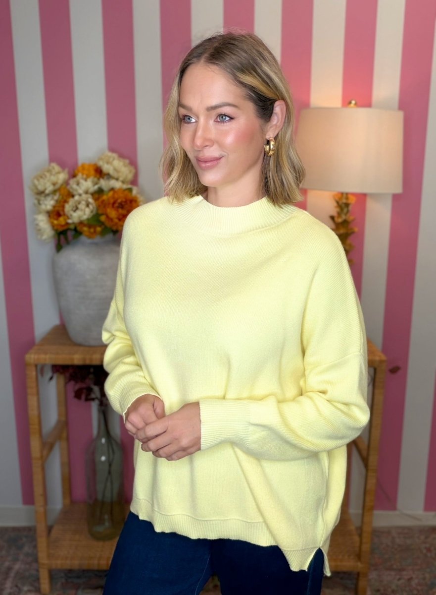 A woman wearing a pale yellow crewneck sweatshirt and dark blue jeans, standing in front of a pink and white striped wall with a lamp and a vase of flowers in the background.