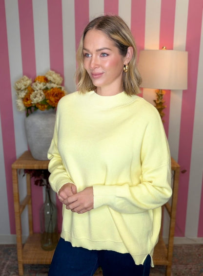 A woman wearing a pale yellow crewneck sweatshirt and dark blue jeans, standing in front of a pink and white striped wall with a lamp and a vase of flowers in the background.