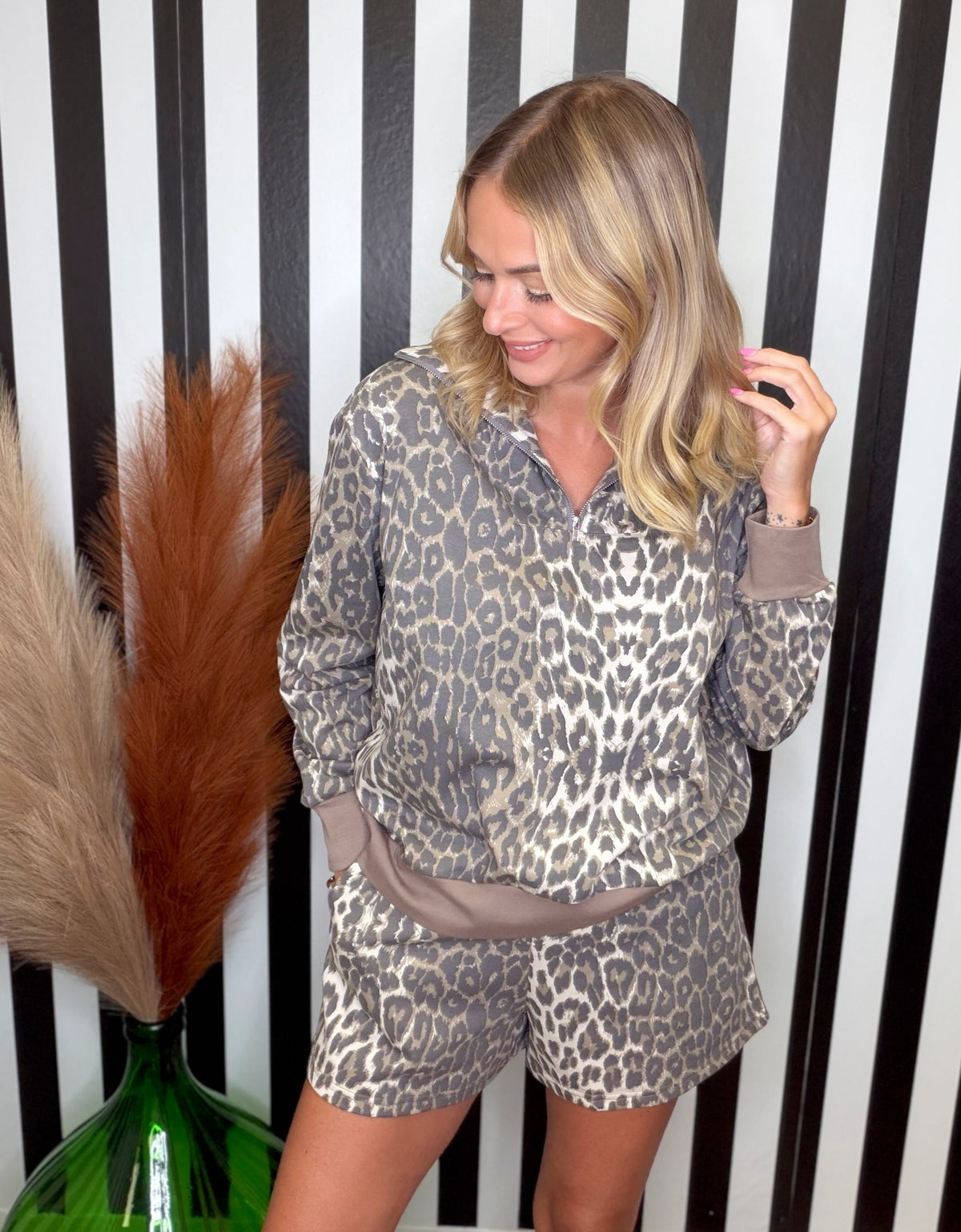 A woman modeling a matching two-piece loungewear set featuring a beige, brown, and black leopard print pattern with solid taupe-brown trim, posed in front of a black and white striped background.