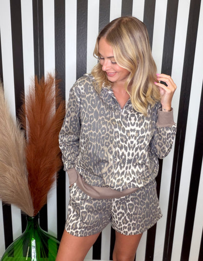 A woman modeling a matching two-piece loungewear set featuring a beige, brown, and black leopard print pattern with solid taupe-brown trim, posed in front of a black and white striped background.