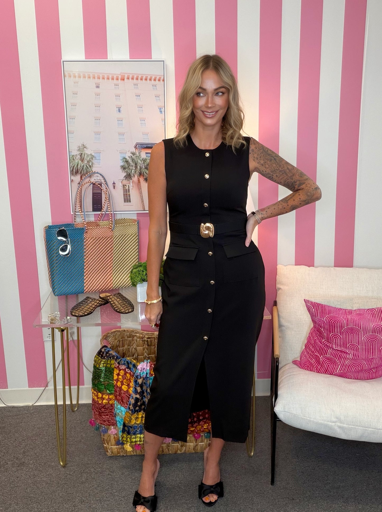 A woman stands indoors wearing a sleeveless black midi dress with a front button placket, waist belt, and front slit. The room has pink-and-white striped walls, a small table with handbags and a colorful quilt.