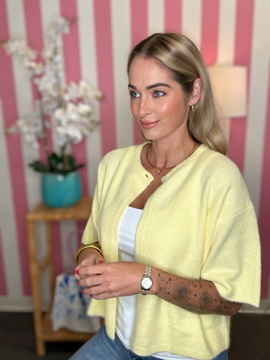 A woman wearing a pale yellow knit cardigan over a white top and blue jeans, standing in front of a pink and white striped wall with a small wooden side table and flowers in the background.