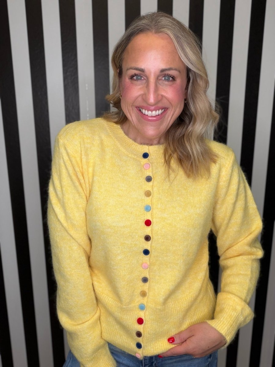A smiling blonde woman wearing a fuzzy yellow knit sweater with a vertical row of colorful buttons, paired with blue jeans, standing against a black and white striped background.