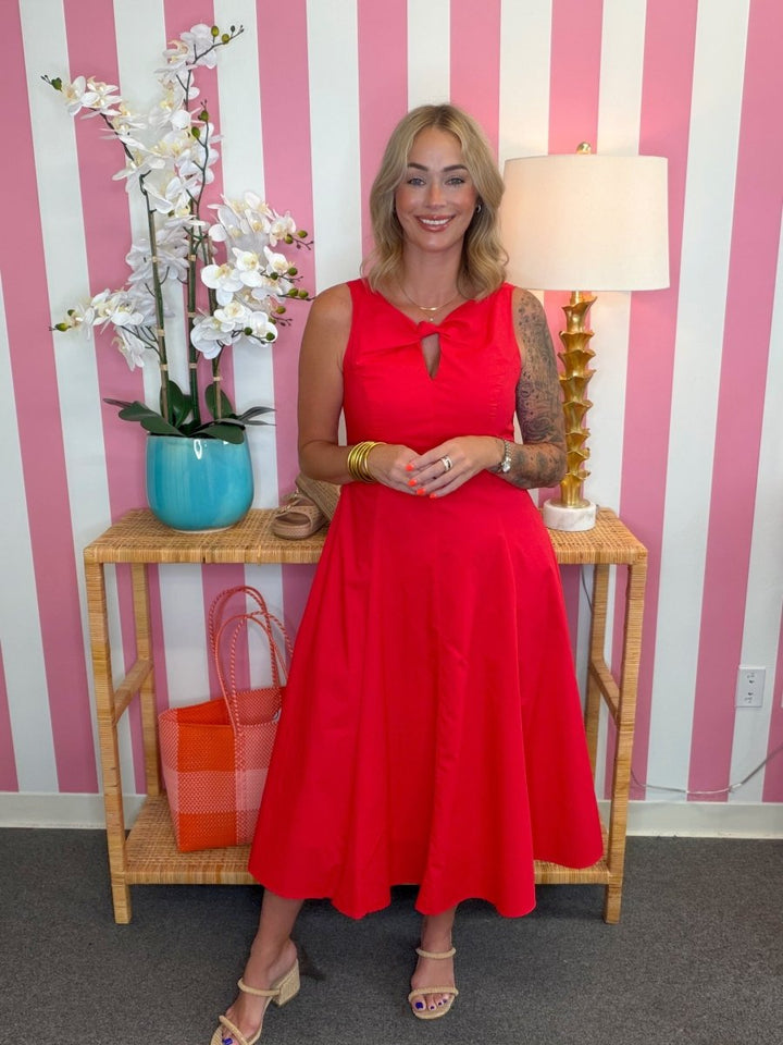 Woman in a red dress standing in front of a pink and white striped wall with a table and lamp.
