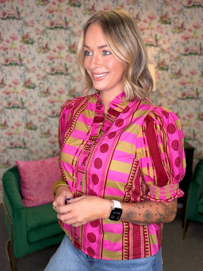 A woman wearing a vibrant pink and olive green patterned blouse with puff sleeves, standing in front of a floral wallpaper background.