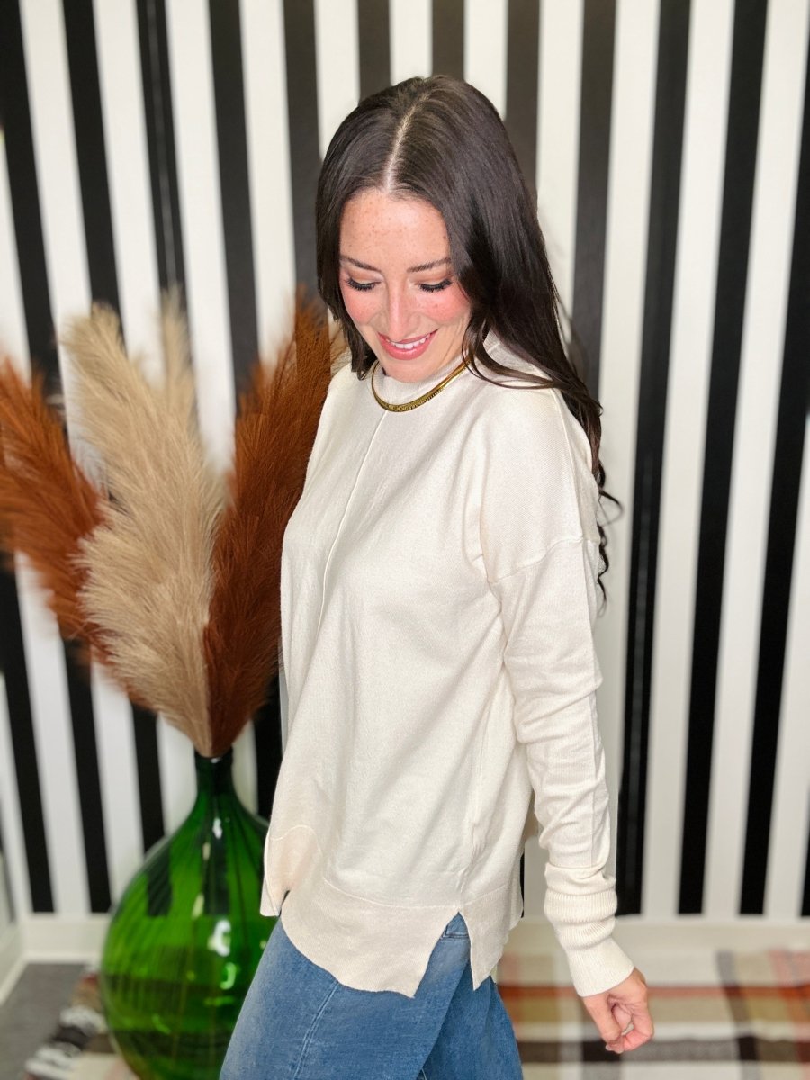 A woman wearing a cream-colored long-sleeve knit top and blue jeans, standing in front of a black and white striped wall with a green vase of pampas grass on the left.
